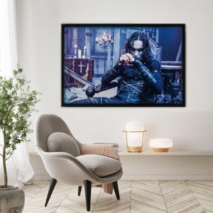 Puede incluir: Impresión enmarcada de una persona vestida de oscuro, sentada en una silla, con un fondo interior gótico. La persona tiene maquillaje oscuro y sostiene un bastón decorativo. La impresión está en un marco negro y cuelga de una pared blanca.