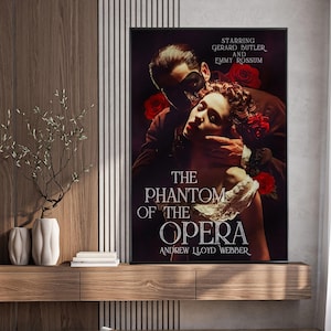 Könnte beinhalten: Gerahmtes Poster für das Musical "Das Phantom der Oper" mit Gerard Butler und Emmy Rossum. Das Kunstwerk zeigt einen Mann mit Maske und eine Frau, mit roten Rosen. Titel und Name des Komponisten sind abgebildet.