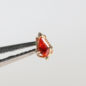 May include: A small, kite-shaped orange gemstone set in a delicate gold-toned metal prong setting. The piece is held by silver-colored metal tweezers, showcasing the faceted surface of the stone and the intricate metalwork of the setting.