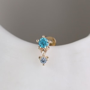 May include: A gold-toned stud earring featuring a prong-set bright blue circular gemstone at the top, connected by a small metal link to a smaller, lighter blue circular gemstone dangling below in a vertical arrangement.