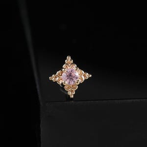 May include: A gold stud earring featuring a pink gemstone at its centre. The earring has a four-pointed star design with intricate detailing. The background is black.