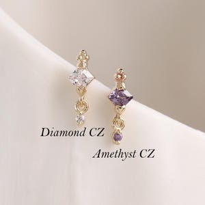 May include: Two gold-tone stud earrings featuring vertical designs. The left earring has a clear diamond CZ stone, and the right earring has a purple amethyst CZ stone. Both earrings include small dangling accents and are labeled Diamond CZ and Amethyst CZ.