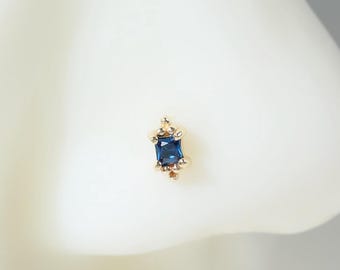 Peut inclure: Un bijou de nez en or orné d'une pierre précieuse rectangulaire bleu profond, montée verticalement. La monture présente de petits détails dorés délicats en haut et en bas de la pierre, offrant une apparence raffinée et symétrique.