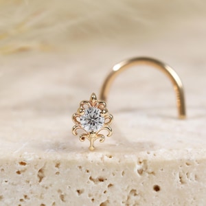 May include: A gold nose stud featuring a curved post and a decorative filigree setting. The centre of the ornament holds a single round clear gemstone. The metalwork displays intricate scroll patterns surrounding the central stone.
