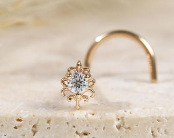 Peut inclure: Un bijou de nez en or avec une tige courbée et un sertissage en filigrane décoratif. Le centre de l'ornement contient une pierre précieuse ronde et transparente. Le métal présente des motifs de volutes complexes autour de la pierre.
