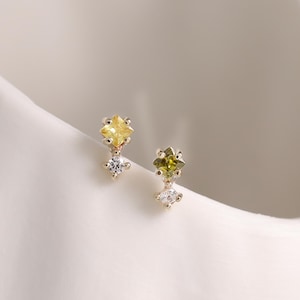 May include: A pair of gold-toned earrings, each featuring a yellow and green gemstone with a small white gemstone below. The earrings are set against a white background, showcasing their delicate design.