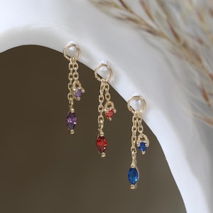 May include: Three gold-toned dangle earrings, each featuring a small hoop attached to two delicate chains. The earrings are adorned with small, faceted marquise and round-cut gemstones in purple, red, and blue, suspended from the gold chains.