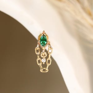 May include: A gold stud earring featuring a marquise-cut green gemstone at the top, connected to two delicate dangling gold chains that form a loop, with a small clear round gemstone suspended in the center of the chain structure.