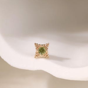 May include: A small, square gold earring featuring an intricate pattern and a central green gemstone. The earring is set against a white, curved backdrop, with a delicate, feathery plant in the upper left corner.