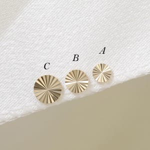 May include: Three circular gold-toned metal studs arranged in a row, labelled A, B, and C from smallest to largest. Each stud features a sunburst pattern with fine, radiating lines etched into the surface, creating a reflective, textured finish.