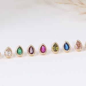 May include: A selection of gold-coloured teardrop-shaped stud earrings, each with a different coloured gemstone. Gemstones include red, purple, blue, clear, green, and pink. The earrings are displayed on a white surface.