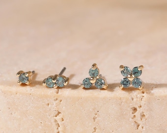 May include: Four gold stud earrings arranged in a row, each featuring small, round, light blue stones. The first earring has one stone, the second has two, the third has three in a triangular cluster, and the fourth has four in a flower shape.