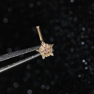 May include: A rose gold nose stud featuring a small, multi-petaled flower design with a central stone. The jewelry piece is held by silver-toned metal tweezers, showcasing the intricate floral metalwork and the delicate, polished finish of the stud.
