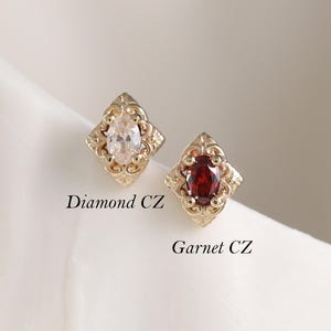 Puede incluir: Dos pendientes de botón dorados con un marco de metal ornamentado en forma de diamante. Uno tiene una piedra CZ de diamante de talla marquesa transparente, el otro una piedra CZ de granate de talla ovalada roja. Etiquetados Diamond CZ y Garnet CZ.