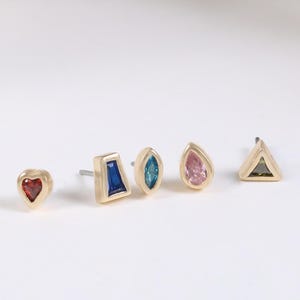 Op de afbeelding: Vijf gouden oorknopjes op een rij, elk met een andere vorm en kleur edelsteen: een rood hartje, een blauwe trapeze, een blauwgroene marquise, een roze druppel en een donkere driehoekige steen in een gouden metalen zetting.