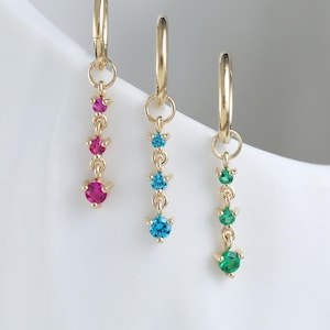 May include: Three gold hoop earrings, each featuring a dangling chain of three small round gemstones. The first earring has pink stones, the second has bright blue stones, and the third has green stones, all set in polished gold metal settings.