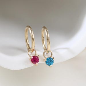 May include: A pair of gold hoop earrings, each featuring a small dangling charm. One charm is set with a round, vibrant red gemstone, while the other charm is set with a round, bright blue gemstone, both held in place by delicate metal prongs.