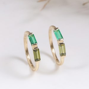 May include: A pair of gold hoop earrings, each featuring two baguette-cut gemstones. One stone on each hoop is a vibrant emerald green, while the other is a lighter, olive-toned green. The hoops are thin, polished gold metal, designed as small circular bands.