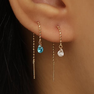 May include: Two gold threader earrings featuring delicate chain designs. One earring displays a small, bright blue teardrop-shaped stone, while the other earring features a clear, white teardrop-shaped stone, both hanging from thin gold chains.