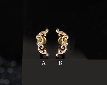 May include: Two gold-toned stud earrings featuring an ornate, curved scrollwork design. Each earring is marked with a letter below it, with the left earring labeled A and the right earring labeled B, showcasing intricate metal detailing.