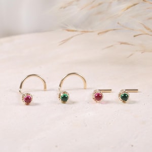 May include: Four gold nose studs arranged in a row. Two studs feature small, round, deep red gemstones, and two studs feature small, round, dark green gemstones. Each gemstone is set in a delicate gold bezel with a thin, curved or straight post.