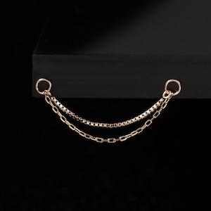 May include: A gold-coloured double chain earring with two small hoops at the top. The chains are different styles, one is a box chain and the other is a cable chain. The earring is displayed against a black background.