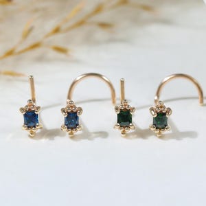 May include: Four gold nose studs arranged in a row. Each stud features a small rectangular gemstone in shades of blue and green, held by four tiny prongs. The metal posts are curved, designed for piercing, with a polished gold finish throughout.