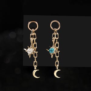 May include: A pair of gold-toned drop earrings featuring a delicate chain link design. Each earring displays a star-shaped charm with a central gemstone, one clear and one teal, finished with a small crescent moon charm at the base.