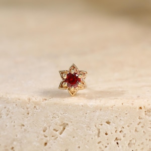 May include: A gold stud earring featuring a six-pointed star-shaped setting with a small, round, deep red gemstone at its center. The metal has a polished finish, and the design is symmetrical with pointed edges surrounding the central stone.