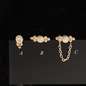 May include: Three gold-toned stud earrings labeled A, B, and C. A features a single round stone with bead accents. B has a central round stone with side bead details. C includes a round stone with a dangling gold chain and bead accents.