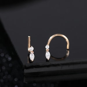 Puede incluir: Dos piercings de nariz dorados, cada uno con pequeñas piedras preciosas claras en forma de lágrima. Uno es una barra recta con dos gemas, el otro un aro curvo con una gema. La joyería se encuentra sobre un fondo oscuro.