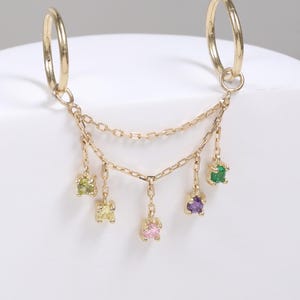 Könnte beinhalten: Ein goldener Doppel-Creolen-Ohrring, der durch zwei zarte Ketten verbunden ist und fünf kleine hängende Edelsteine in Hellgrün, Gelb, Rosa, Violett und Dunkelgrün aufweist. Das Schmuckstück besteht aus feinen Metallgliedern und gefassten Steinen.