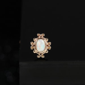 Peut inclure: Une boucle d'oreille à clou en or rose ornée d'une opale blanche ovale en cabochon, sertie dans une monture métallique décorative et ornée de détails en volutes complexes. La pierre centrale présente des reflets irisés sur le métal poli.