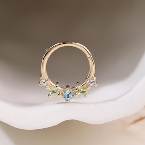 Puede incluir: Un aro de tabique dorado con cierre de bisagra. El aro está adornado con una fila de piedras preciosas, que incluyen piedras de color azul claro, verde y transparente. Las piedras preciosas están engastadas en un patrón decorativo.