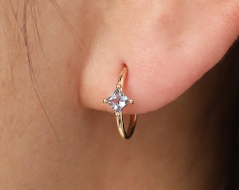 May include: A small gold hoop earring featuring a single, square-cut, light blue gemstone set in a four-prong setting. The earring is positioned in the earlobe, showcasing the polished gold band and the clear, pale blue stone centered on the hoop.