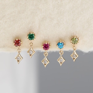Puede incluir: Cinco pendientes colgantes de oro dispuestos en fila. Cada pendiente presenta una piedra preciosa redonda facetada en rojo, verde, rosa, azul o verde claro, unida por un pequeño eslabón de oro a un dije en forma de rombo con una piedra.