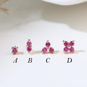 以下が含まれることがあります： ピンク色の宝石が付いた4つのゴールドトーンスタッドピアス。ピアスは一列に並んでおり、A、B、C、Dとラベル付けされています。各イヤリングは、単一の石、石の垂直列、三角形の配置、4石の花など、さまざまなデザインが特徴です。