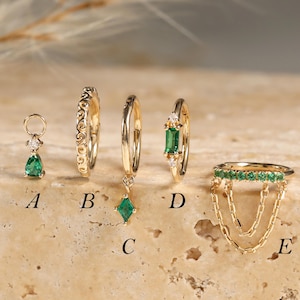 May include: Five gold jewelry pieces labeled A through E. A is a pear-cut green stone charm. B is a textured hoop. C is a hoop with a diamond-shaped green stone charm. D is a hoop with a baguette-cut green stone. E is a ring with green stones and chains.