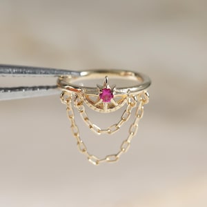 May include: A gold hoop ring featuring a central star-shaped setting with a small round pink gemstone. Two delicate gold chains drape from the sides of the star, connecting to the bottom of the hoop to create a layered, dangling effect.