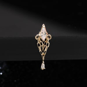 Puede incluir: Un pendiente de color dorado con un diseño geométrico. El pendiente presenta una piedra transparente en forma de diamante en la parte superior, conectada a una cadena que conduce a una piedra transparente en forma de lágrima. El pendiente está sobre un fondo negro.