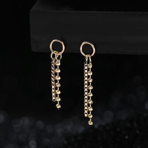 May include: A pair of gold-toned earrings featuring a circular hoop top connected to two dangling chains. One chain is a box link style, while the other is a beaded chain. The earrings have a metallic finish and hang vertically from a dark surface.