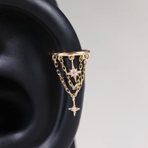 May include: A gold-toned ear cuff featuring a horizontal band connected to two dangling gold chains. The chains are adorned with two four-pointed star charms, each set with a small, light pink stone at its centre, creating a layered design.