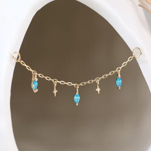 Puede incluir: Una delicada cadena dorada con pequeños abalorios. Los abalorios incluyen tres cuentas en forma de lágrima de color azul claro y dos pequeños abalorios en forma de estrella. La cadena se muestra sobre un fondo blanco.