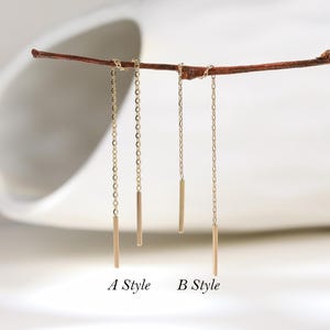 Puede incluir: Dos pares de pendientes de hilo dorados colgados de una fina rama marrón. Los pendientes A Style presentan una cadena más larga en un lado, mientras que los B Style tienen una cadena más corta en un lado. Ambos terminan en una barra metálica.