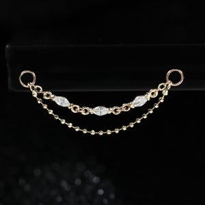 May include: A gold-toned jewellery connector chain featuring two delicate strands. The upper strand is adorned with three marquise-cut clear stones, while the lower strand consists of a fine beaded chain. Both ends terminate in small circular jump rings.