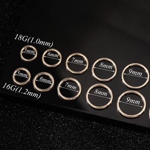 May include: A collection of gold-colored hoop earrings in various sizes, ranging from 5mm to 10mm, and gauges 18G (1.0mm) and 16G (1.2mm). The earrings are arranged on a dark, reflective surface, with size labels clearly visible.