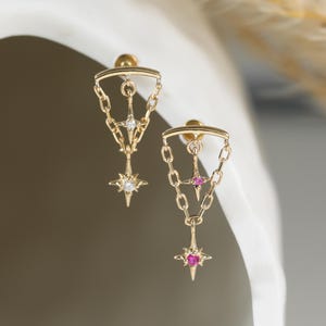 May include: A pair of gold dangle earrings featuring a triangular chain design. One earring is accented with a clear stone at the center and bottom, while the other earring features a vibrant pink stone at the center and bottom star-shaped charm.