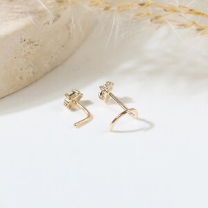 May include: Two gold-toned nose studs featuring a small, dark, rectangular gemstone set in a prong setting. Each stud has a curved L-shaped post designed for secure placement. The metal has a polished finish, reflecting light on the surface.