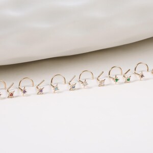 May include: A collection of gold-colored nose rings, each featuring a small star-shaped charm with a different colored gemstone. The gemstones include red, orange, purple, light blue, clear, and green. The nose rings are displayed on a white surface.