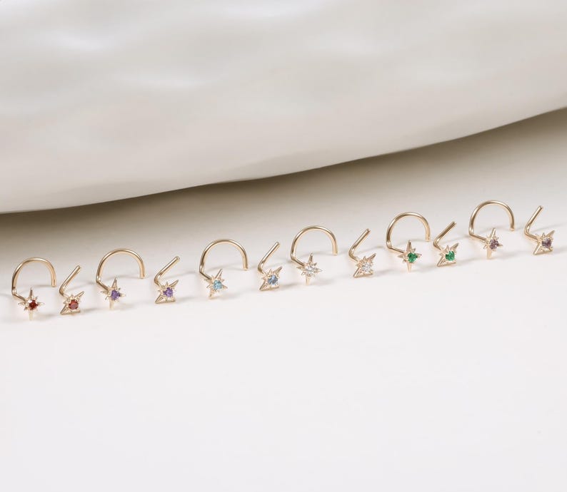 May include: A collection of gold-colored nose rings, each featuring a small star-shaped charm with a different colored gemstone. The gemstones include red, orange, purple, light blue, clear, and green. The nose rings are displayed on a white surface.